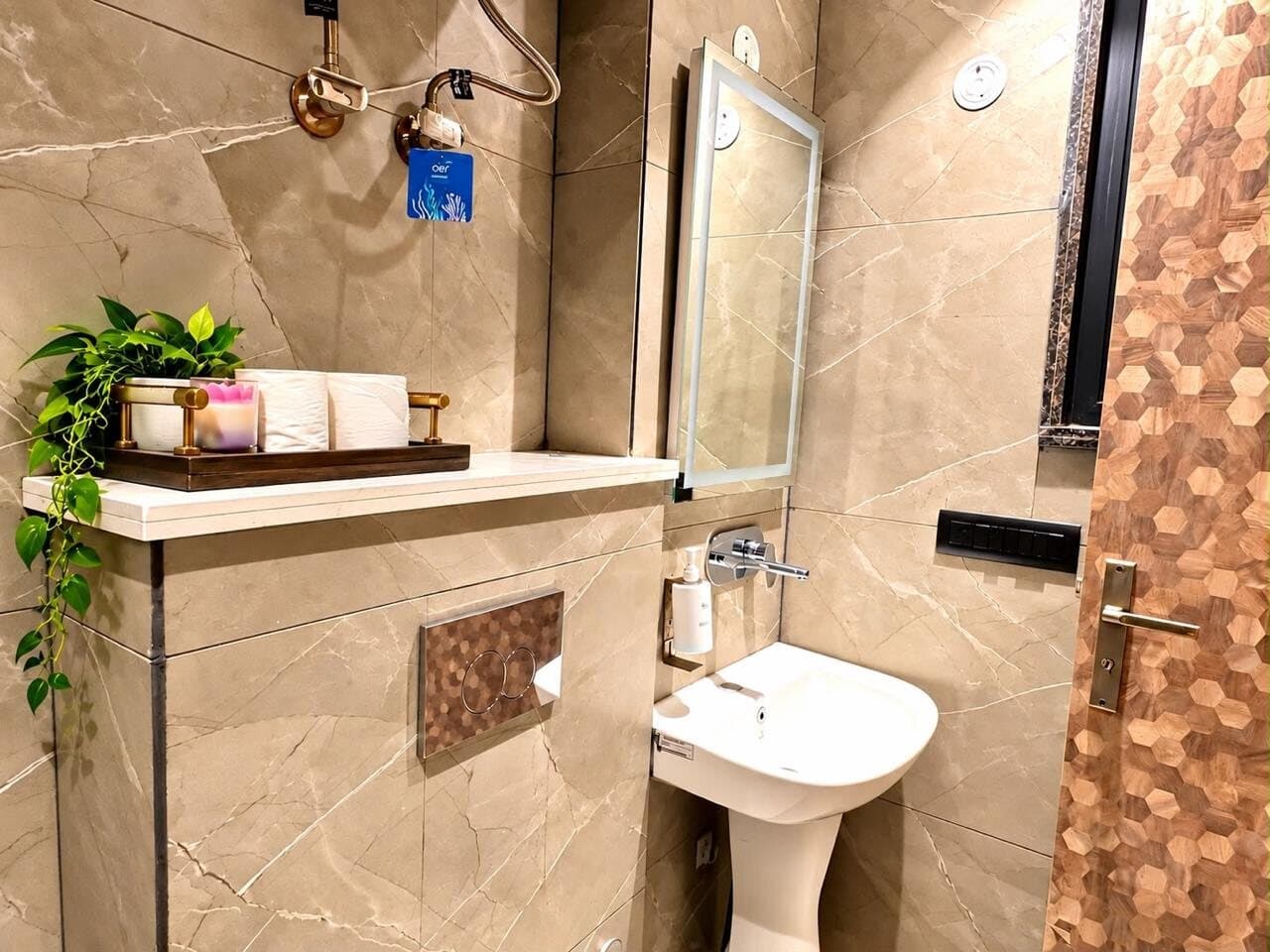 Bathroom with warm marble-look tiles, a backlit mirror, pedestal basin, and a ledge styled with plants and amenities