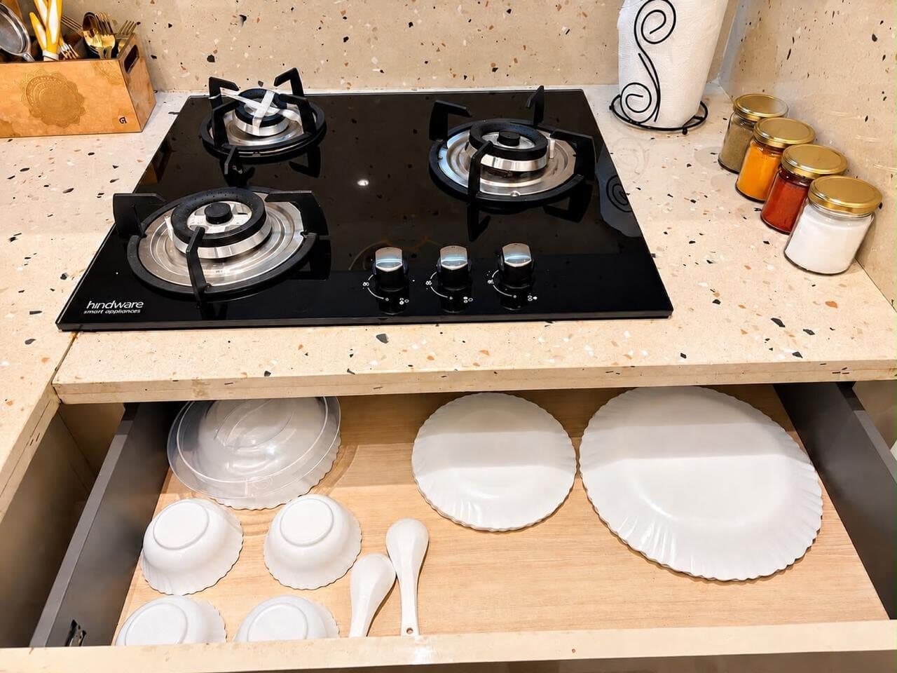 Glass gas hob above a drawer of white dinnerware, with spice jars on the terrazzo counter